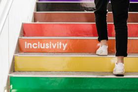 Photo of a pair of legs, dressed in black jeans with white runners, climbing rainbow coloured stairs, with the word inclusivity in white against the orange step.